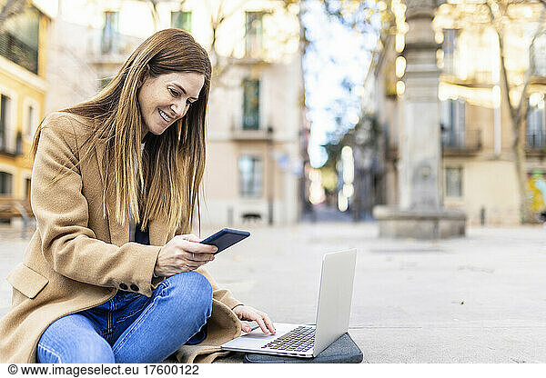 Smiling woman with laptop surfing net through mobile phone sitting on city street