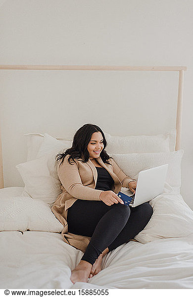 Smiling woman with long dark hair sitting on bed  looking at laptop computer.