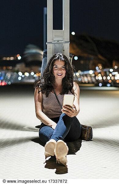 Smiling woman with mobile phone sitting on floor at pier during night