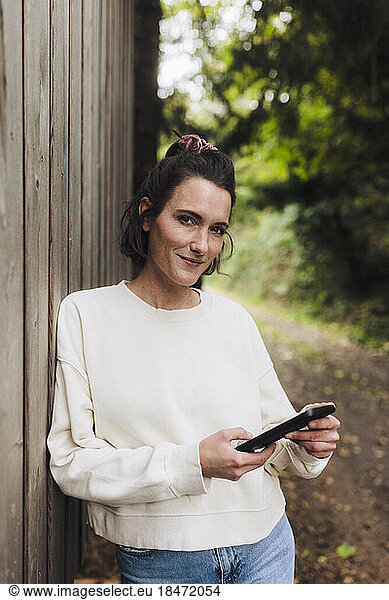 Smiling woman with mobile phone standing by wooden wall