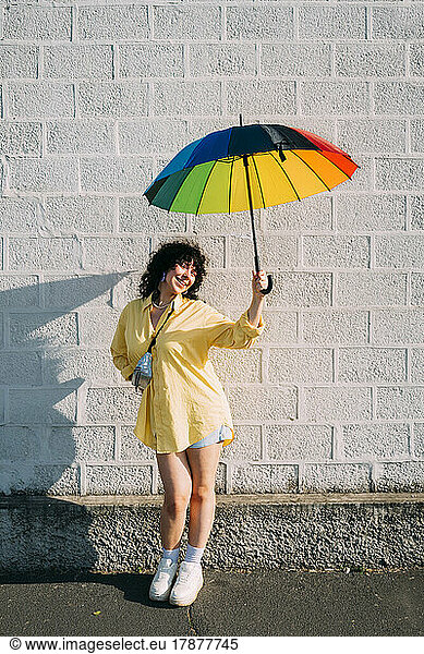 Smiling woman with rainbow umbrella posing in front of wall