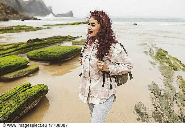 Smiling woman with red hair wearing backpack standing at beach