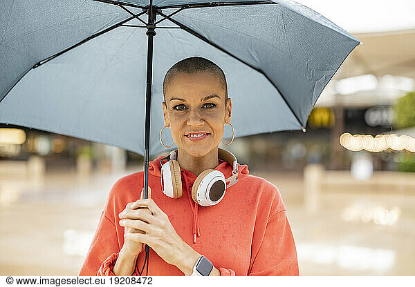 Smiling woman with shaved head holding umbrella in city
