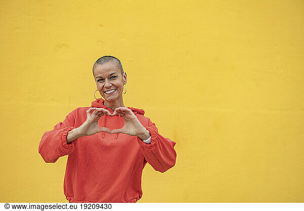 Smiling woman with shaved head making heart shape in front of yellow wall