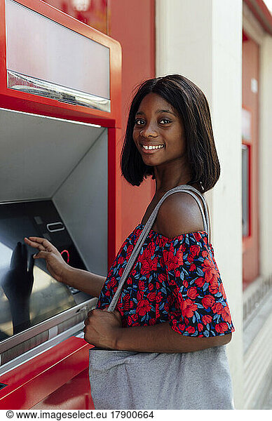 Smiling woman with shoulder bag standing at ATM