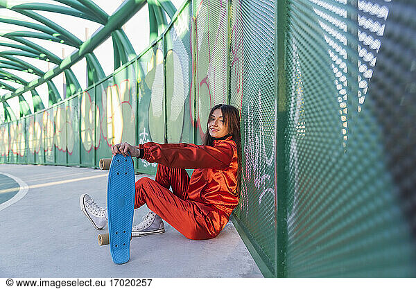 Smiling woman with skateboard resting while sitting on bridge