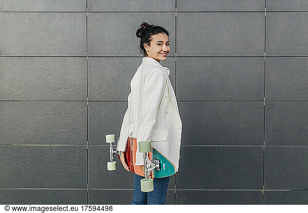 Smiling woman with skateboard standing by wall
