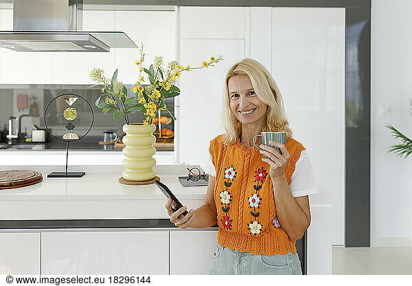 Smiling woman with smart phone and tea cup standing in kitchen
