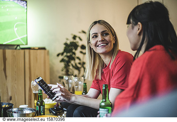 Smiling woman with smart phone looking at friend while watching soccer at home