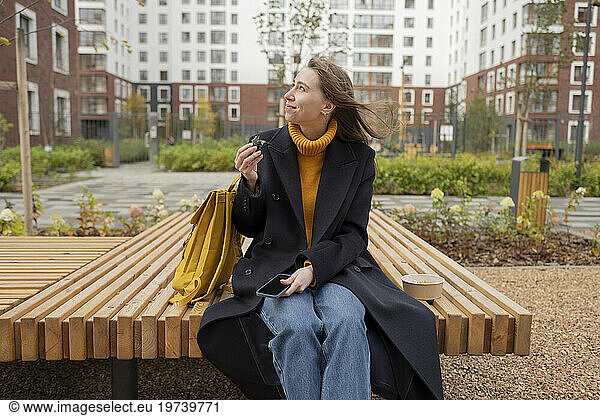 Smiling woman with smart phone sitting on bench and eating lunch at park