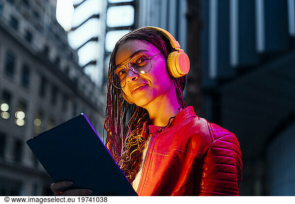 Smiling woman with tablet PC listening music on headphones at dusk