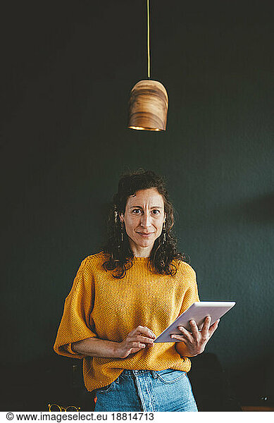 Smiling woman with tablet PC standing under pendant light in front of black wall