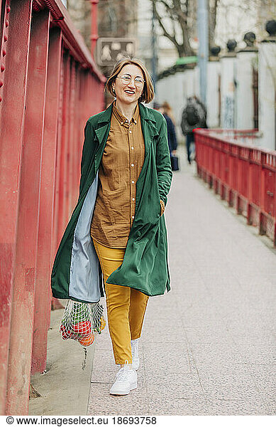 Smiling woman with vegetables in mesh bag walking on footpath