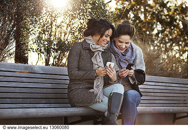 Smiling women using smart phones while sitting on bench