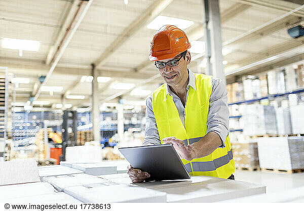 Smiling worker using tablet PC leaning on box in warehouse