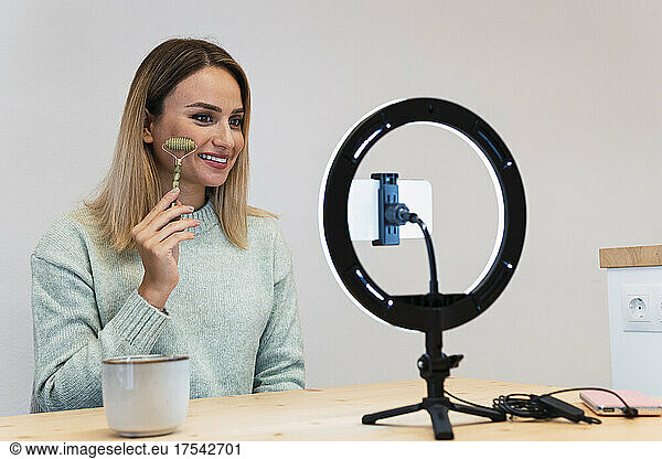 Smiling young beautician making tutorial with jade roller in front of ring light