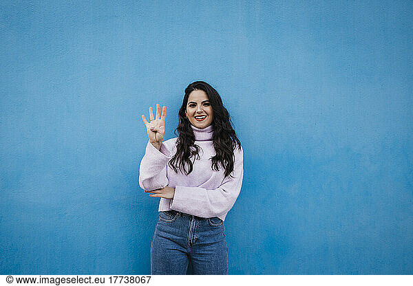 Smiling young beautiful woman showing number 4 in front of blue wall