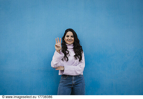 Smiling young beautiful woman showing number 3 in front of blue wall