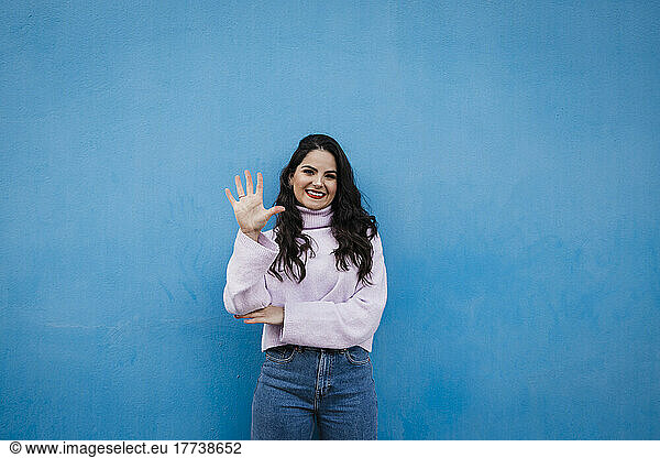 Smiling young beautiful woman showing number 5 in front of blue wall