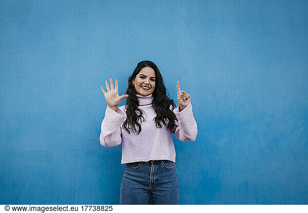Smiling young beautiful woman showing number 6 in front of blue wall