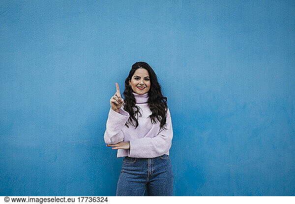 Smiling young beautiful woman showing number one with index finger in front of blue wall