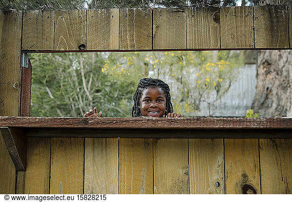 Smiling young black girl with long braids looking through wood fence