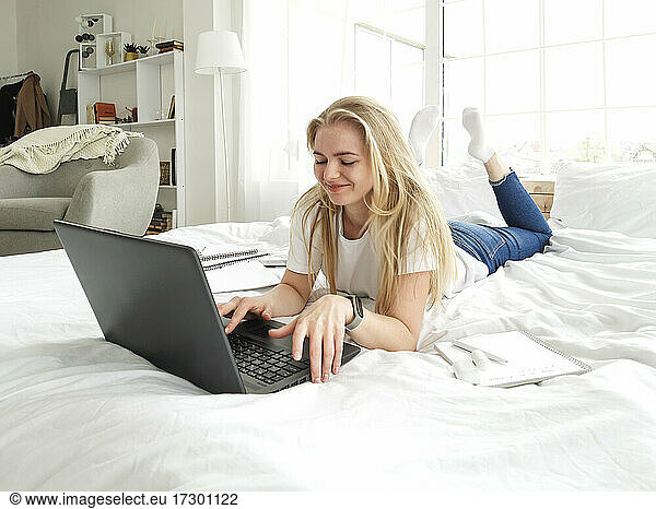 smiling young blond girl typing on laptop lying in the white bed