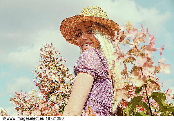Smiling young blonde woman in a straw hat near hydrangea flowers