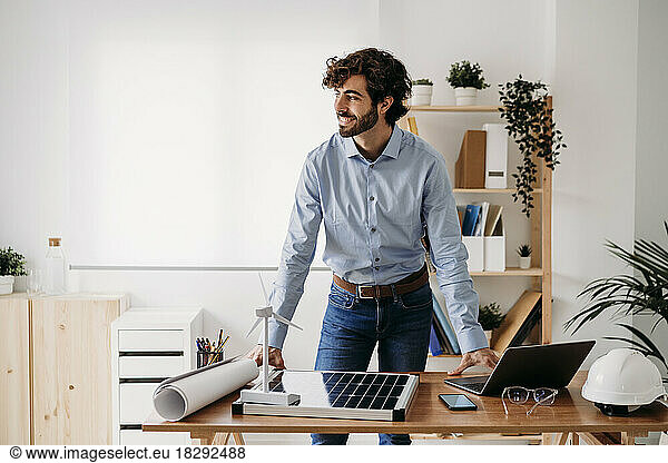 Smiling young businessman with wind turbine and solar panels at desk in office