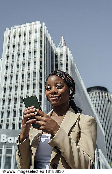 Smiling young businesswoman holding smart phone in front of building