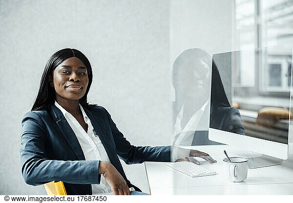 Smiling young businesswoman sitting with desktop PC at desk seen through glass