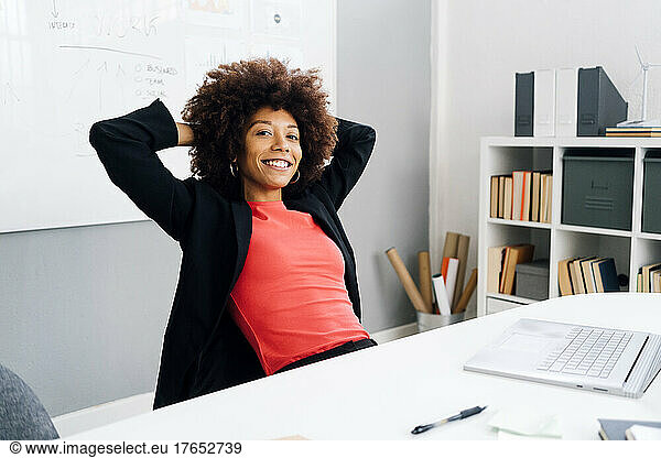 Smiling young businesswoman with hands behind head sitting at desk in office