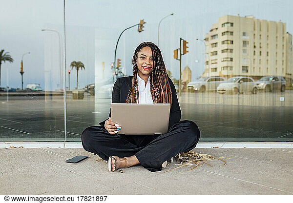 Smiling young businesswoman with laptop sitting in front of glass wall