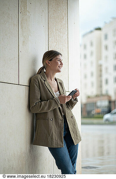 Smiling young businesswoman with mobile phone in front of wall