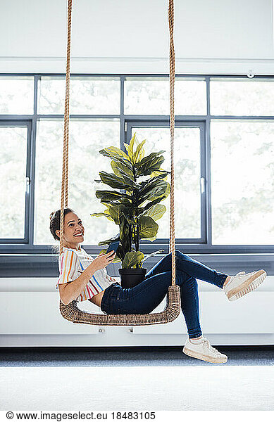 Smiling young businesswoman with potted plant on swing at office