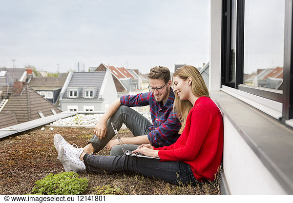 Smiling young couple sitting on roof of city apartment using laptop