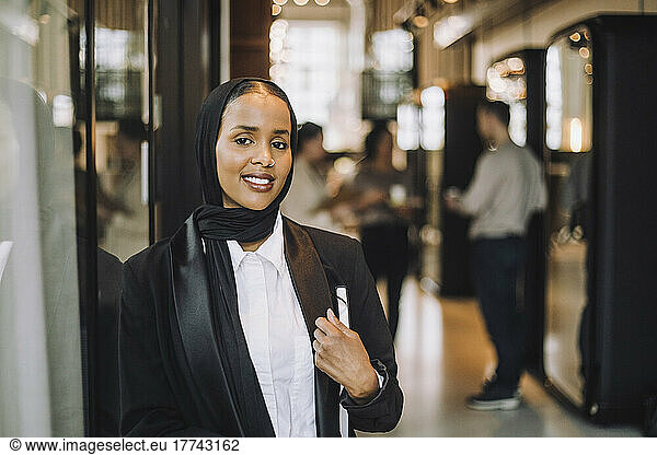 Smiling young entrepreneur wearing headscarf holding diary while standing in office
