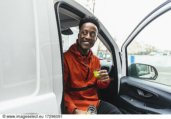 Smiling young essential service man looking away while sitting with drink in delivery van