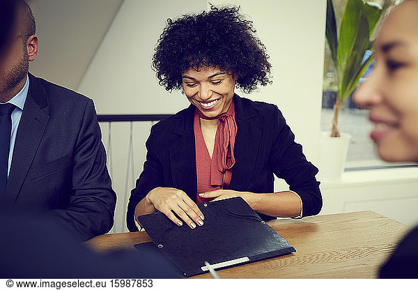 Smiling young female business lawyer holding file while sitting with colleagues at conference table in office during mee