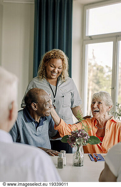 Smiling young female caregiver standing by happy senior women and men at dining table in nursing home