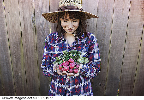 Smiling young female farmer holding radishes while standing against wooden fence