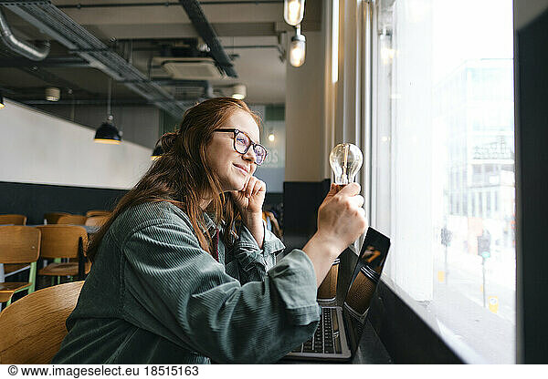 Smiling young freelancer looking at light bulb sitting by window in cafe
