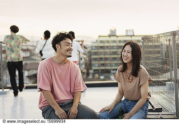 Smiling young Japanese man and woman sitting on a rooftop in an urban setting.