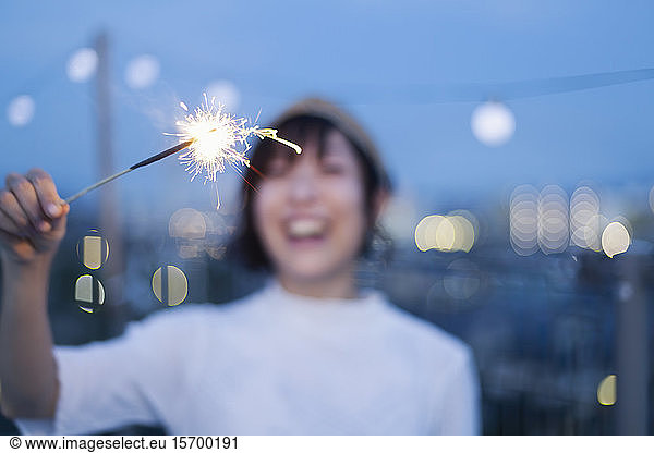 Smiling young Japanese woman holding sparkler on a rooftop in an urban setting.