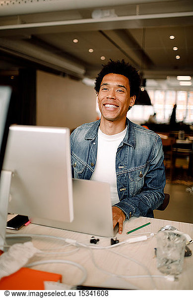 Smiling young male employee using laptop while looking away at desk in office