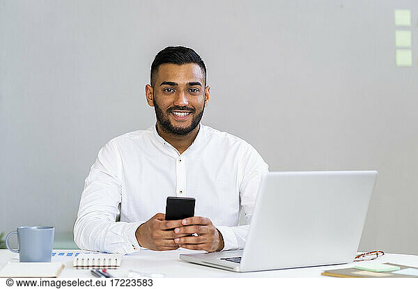 Smiling young male entrepreneur with smart phone by laptop sitting at desk in office