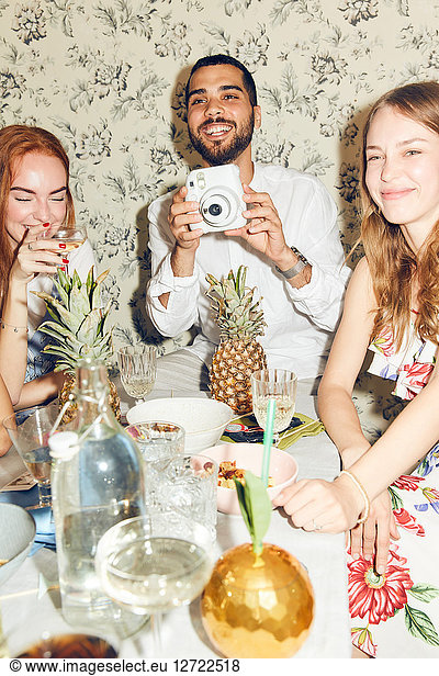 Smiling young man holding camera while sitting amidst female friends at home during dinner party