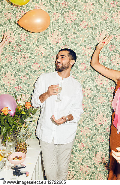 Smiling young man holding drink while standing by woman playing with balloons against wallpaper at home during dinner pa
