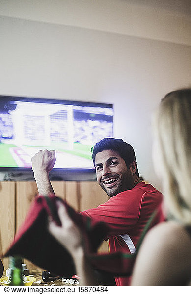 Smiling young man looking at female friend while watching soccer match at home
