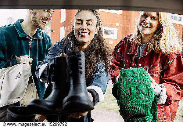 Smiling young man looking at female friends loading shoes and sweater in car trunk